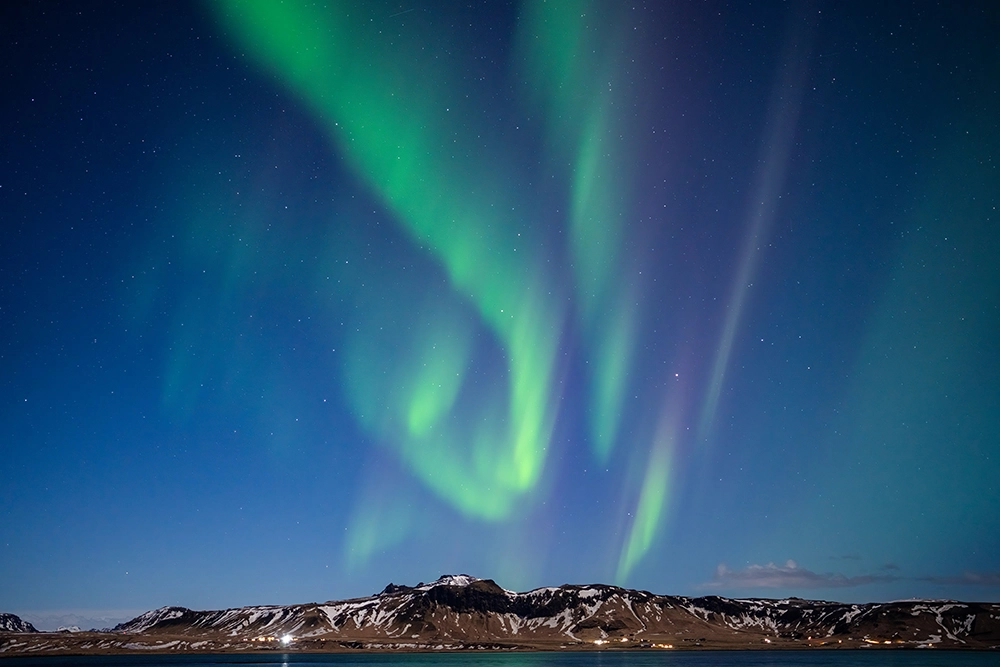 A aurora boreal fotografada com a lente Tamron 35-150 mm F2-2.8, mostrando padrões de aurora verde vibrante acima de uma cadeia de montanhas nevadas em um céu noturno claro.