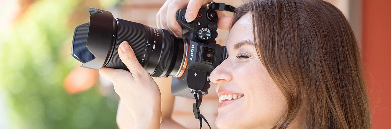 Mujer sonriendo mientras sostiene una cámara sin espejo con un objetivo acoplado bajo la brillante luz del sol.
