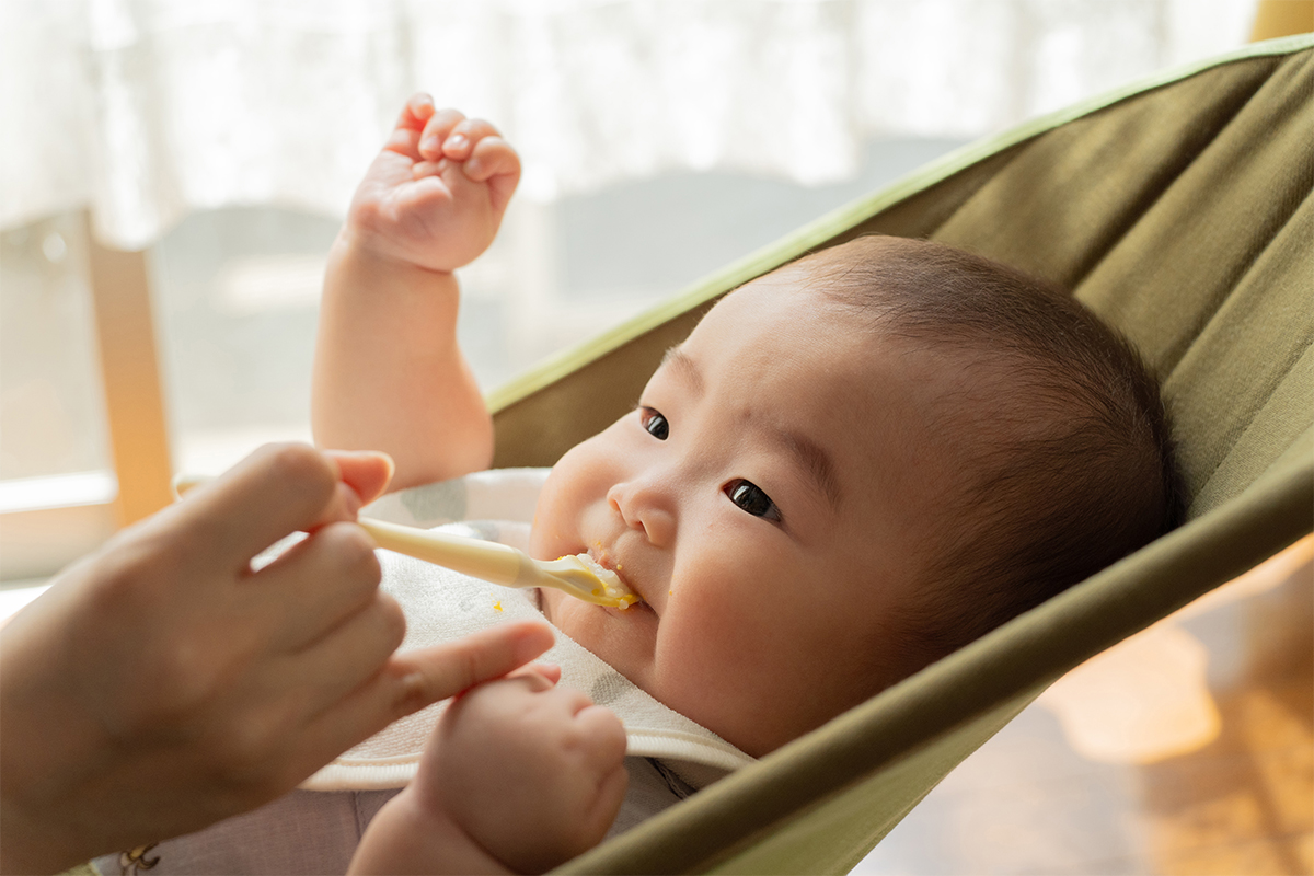 Close-up de um bebê deitado em um assento tipo rede sendo gentilmente alimentado com uma colher sob a luz natural suave de uma janela.