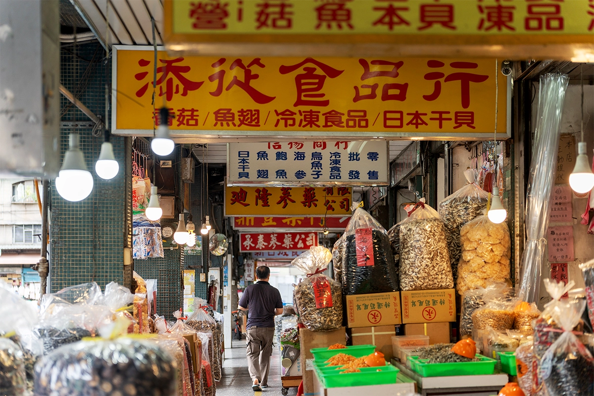 Un viajero caminando por un animado mercado al aire libre lleno de luces colgantes, productos secos y letreros coloridos, capturado para mostrar las historias cotidianas de los viajes.