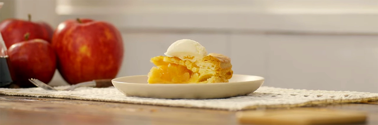 Hero image of a slice of apple pie topped with ice cream on a plate, with apples in the background and soft cinematic bokeh.