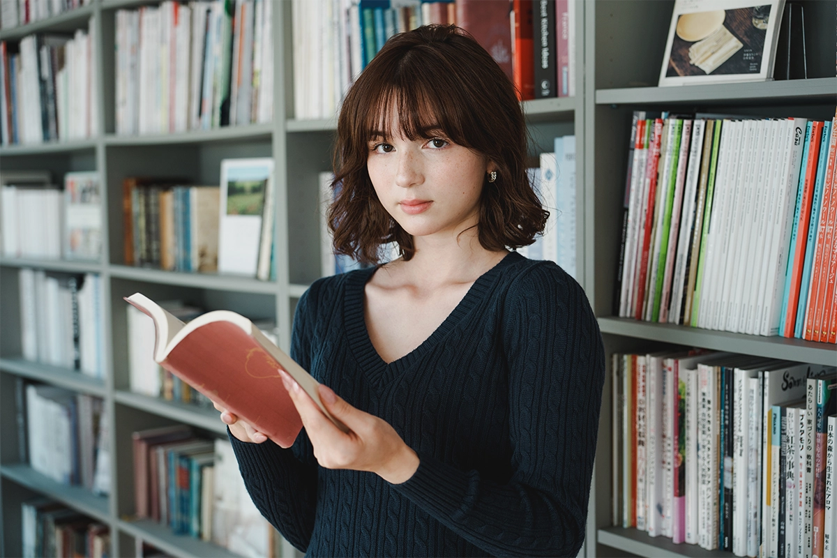Young woman holding a book while standing in front of a bookshelf, photographed in soft natural light for an indoor portrait.