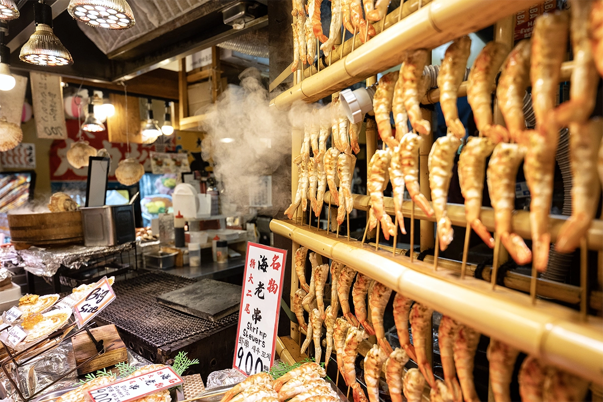 Brochetas de camarones al vapor en un concurrido mercado callejero de comida en Japón, fotografiadas con un lente de viaje Sony E-mount para fotografía cultural y de viajes con poca luz.