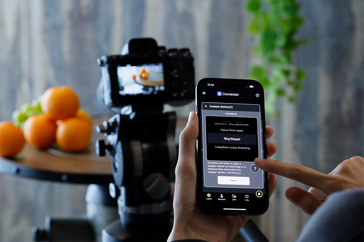 Smartphone exibindo o recurso TAMRON Lens Utility™ Ver. 5.0 Ring Stopper conectado sem fio a uma lente Tamron usando TAMRON-LINK™ durante uma filmagem de vídeo de mesa.