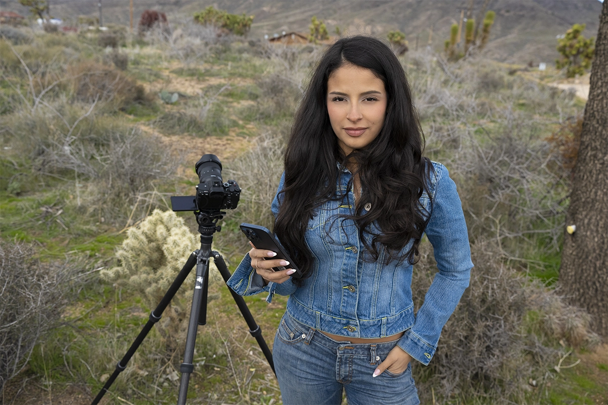 Photographe utilisant TAMRON Lens Utility Mobile connecté sans fil à un smartphone avec TAMRON-LINK à côté d'un appareil photo sans miroir monté sur trépied dans un paysage désertique, démontrant le contrôle sans fil de l'objectif pour la préparation de la photographie du ciel nocturne.