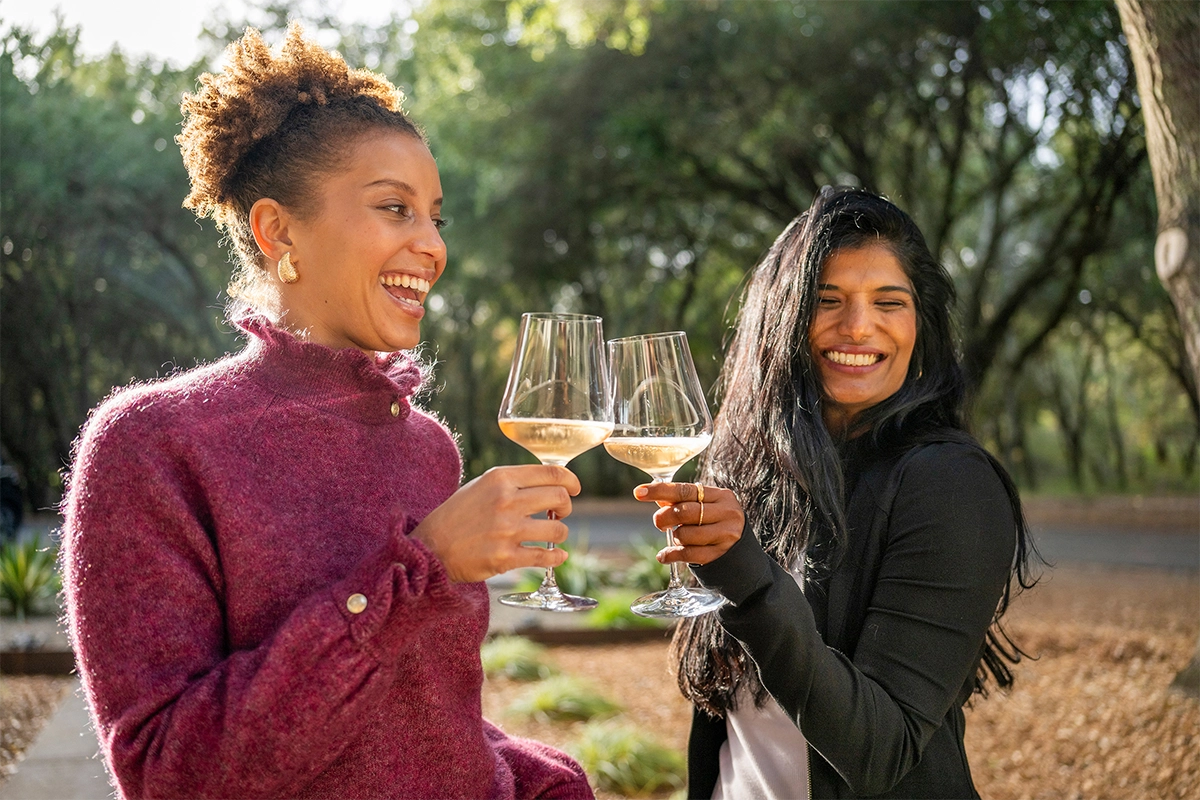 Duas mulheres sorrindo e brindando com taças de vinho branco ao ar livre, sob luz natural quente e cercadas por árvores.