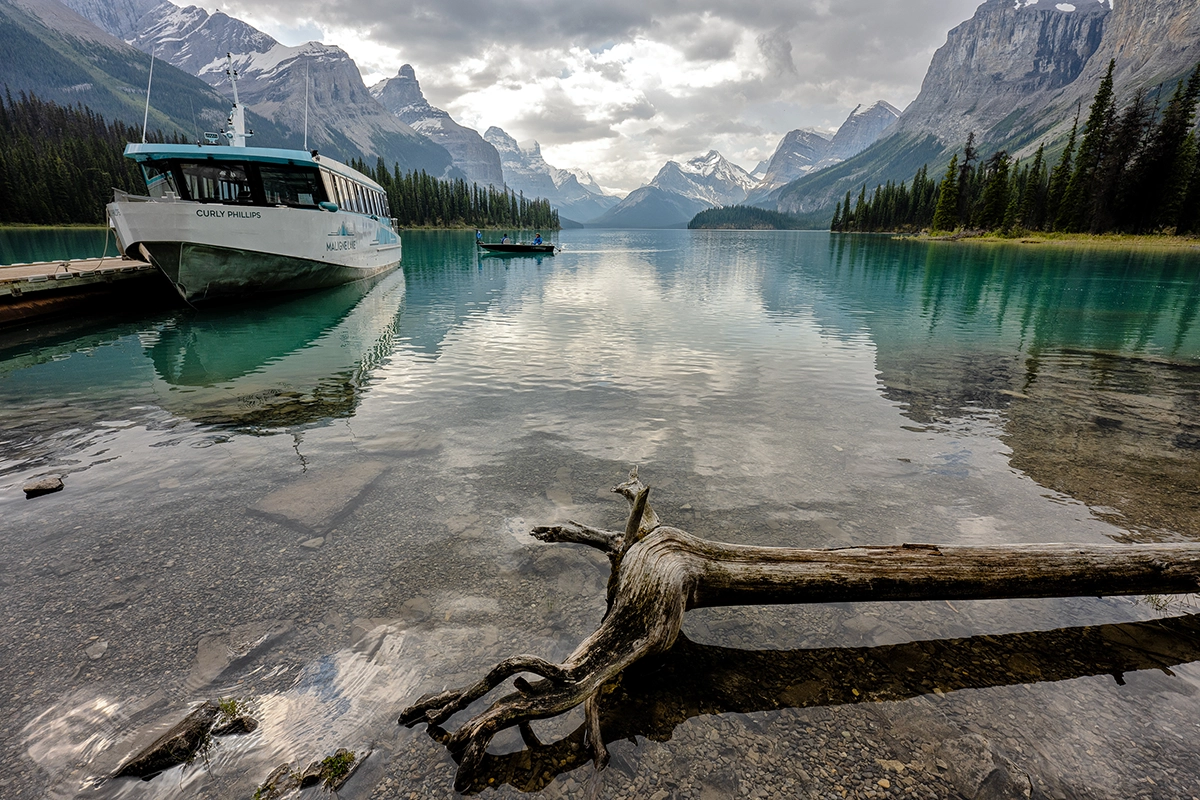 Ultra wide-angle landscape photo of a mountain lake with a driftwood foreground and a boat on the water.