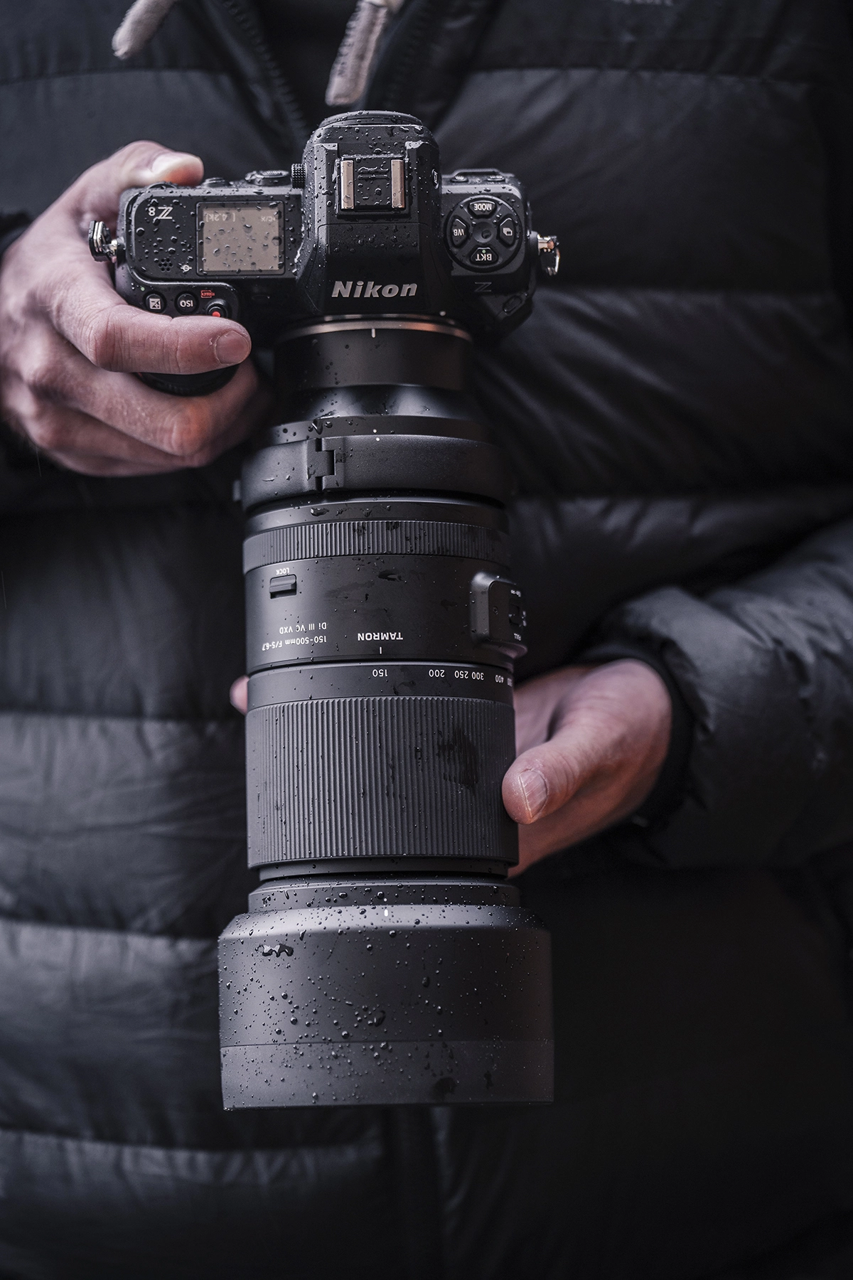 Close-up of a camera and telephoto zoom lens covered in raindrops, illustrating weather sealing and outdoor durability.