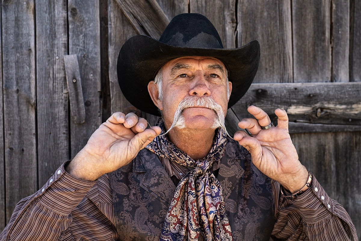 Cowboy com um grande bigode posando para um retrato em um rancho rústico, fotografado com a lente Tamron 35-100 mm F/2.8 Di III VXD.