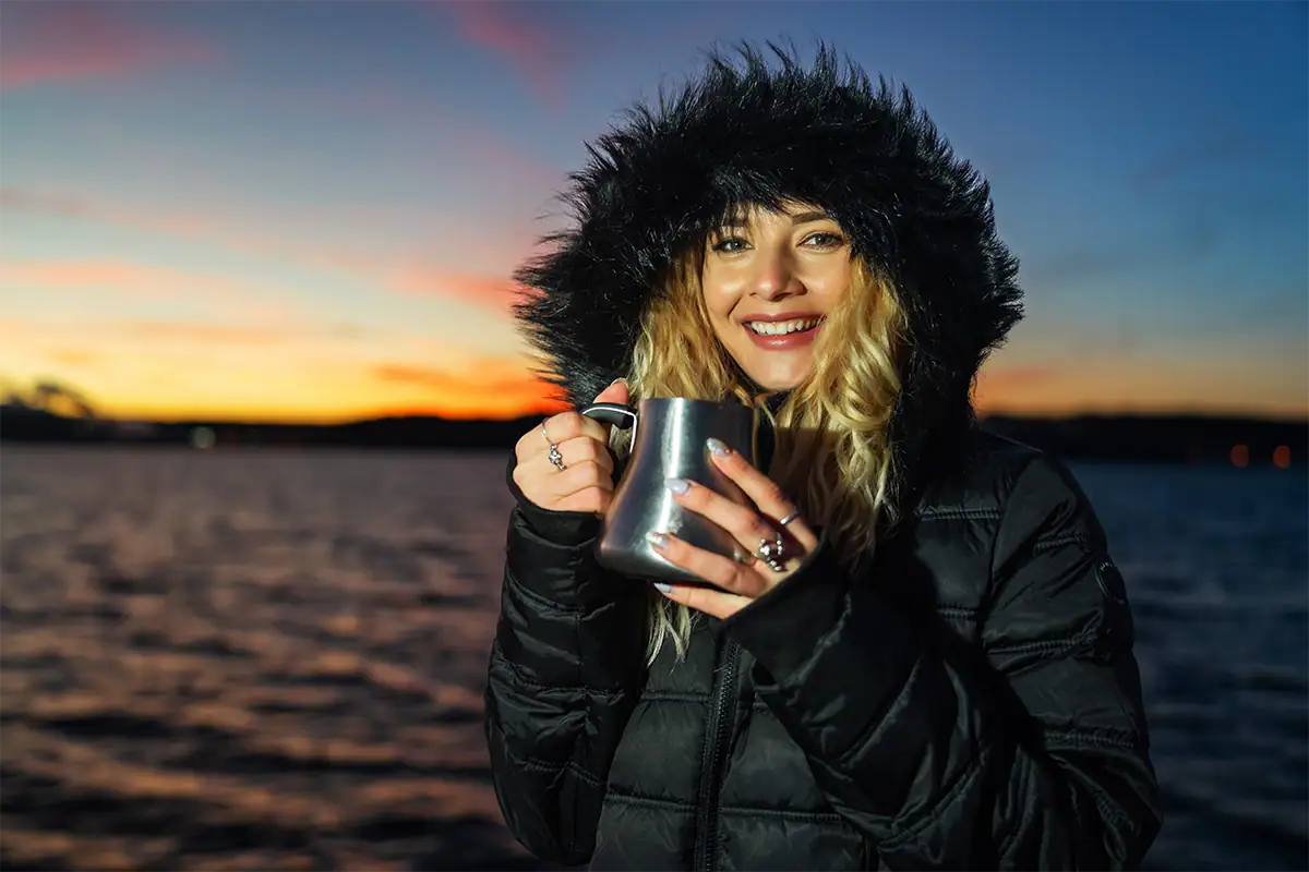 Retrato iluminado con luz artificial durante la hora azul, en el que aparece una mujer con una capucha forrada de piel sosteniendo una taza de metal junto al agua.
