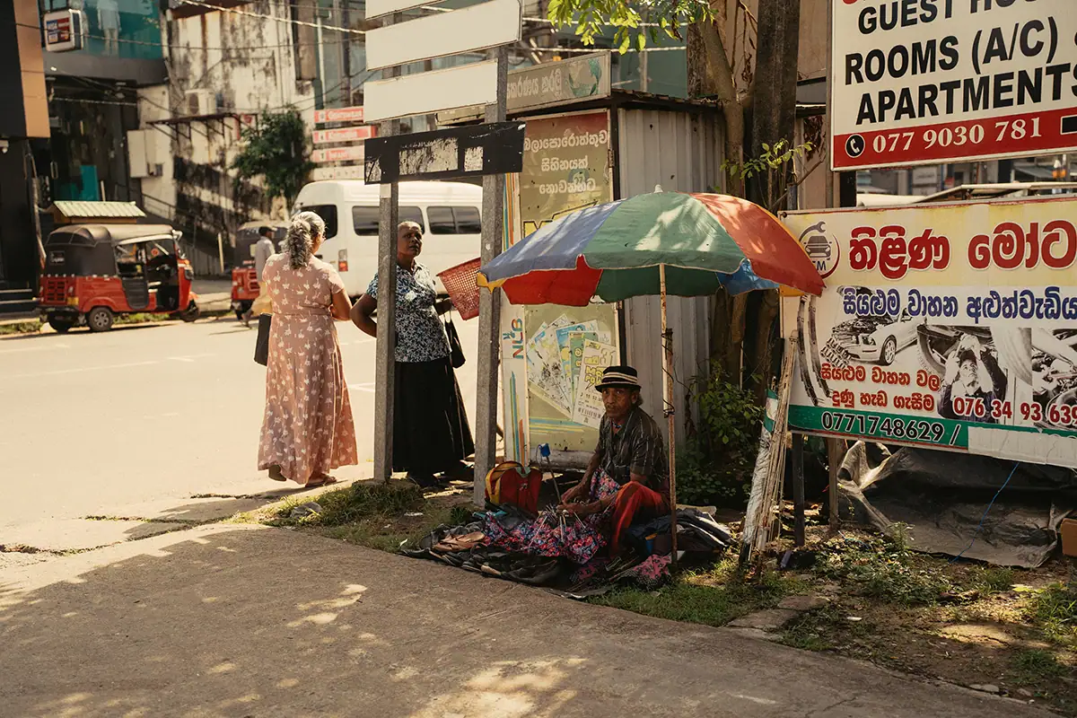 A man fixing umbrellas by the side of the road, with a woman waiting for hers to be repaired.