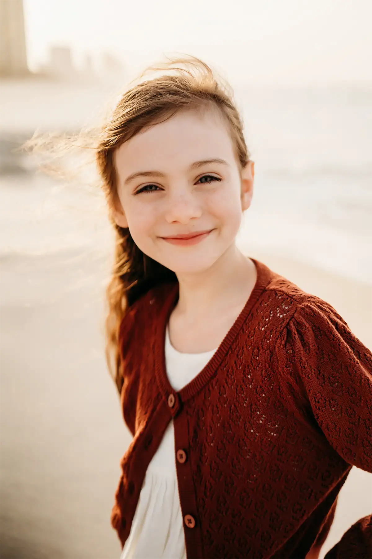 Una niña con un suéter rojo, fotografiada en la playa bajo la suave luz de la madrugada, con reflejos delicados y tonos de piel naturales. 