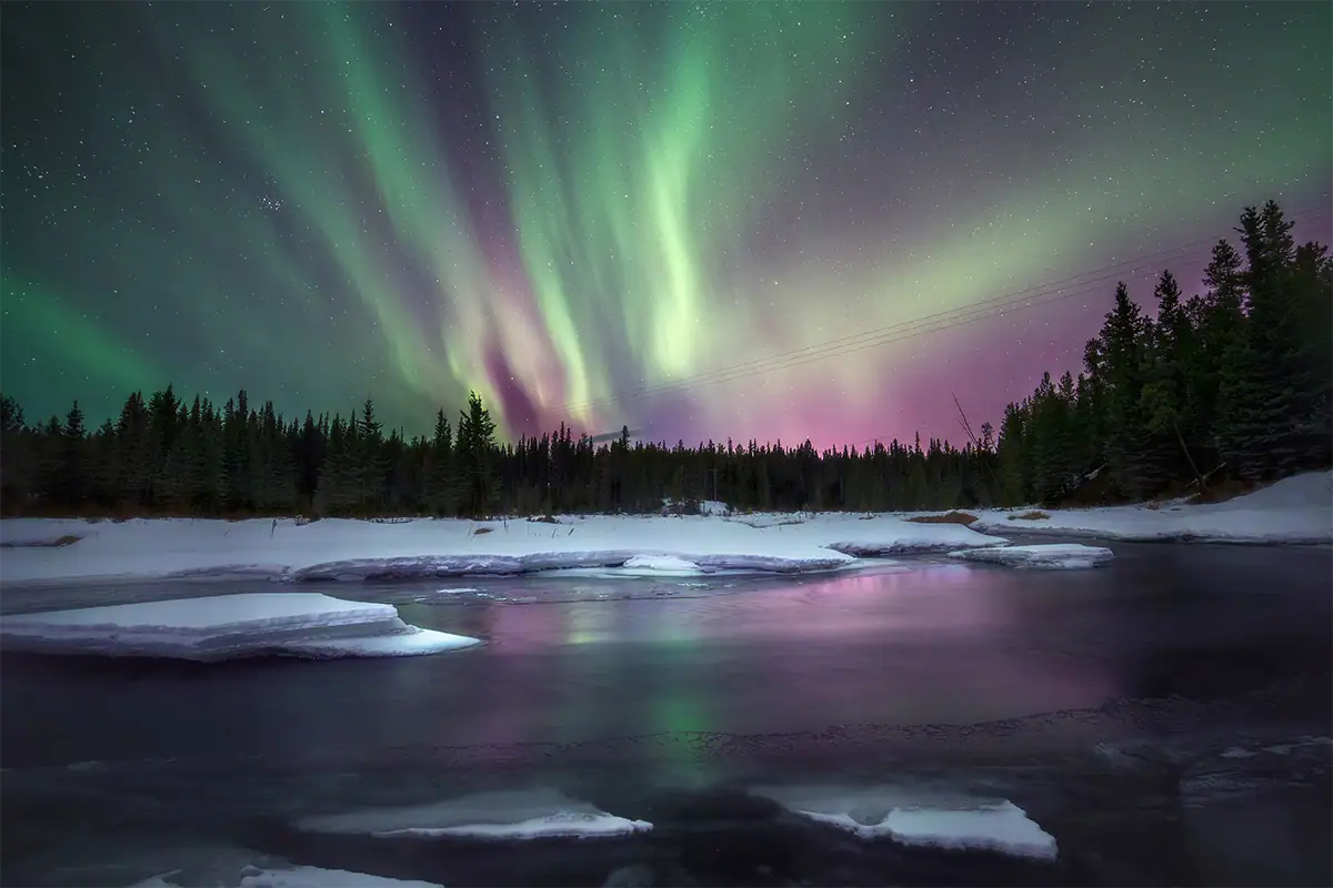 Wide-angle night landscape of the aurora borealis over a snowy river and forest.
