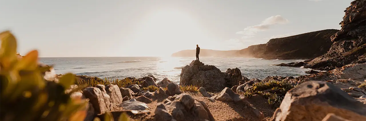 Person standing on rocks overlooking the ocean in a wide-angle coastal landscape.