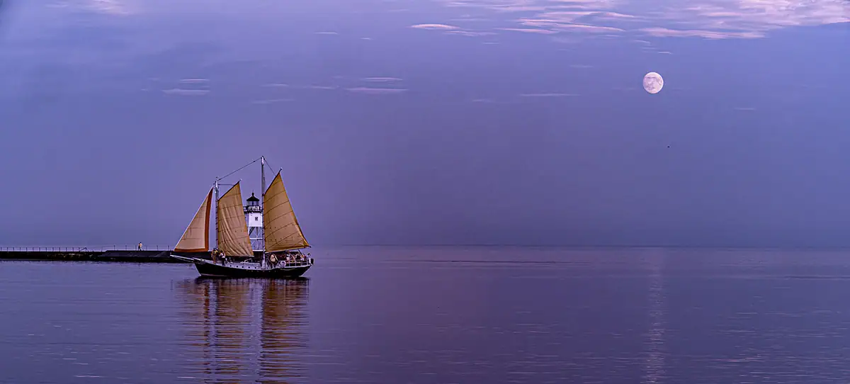 The harbor in Grand Marais, Minnesota, with a lighthouse peeking out from between the sails of the Hjordis schooner as the full moon is rising