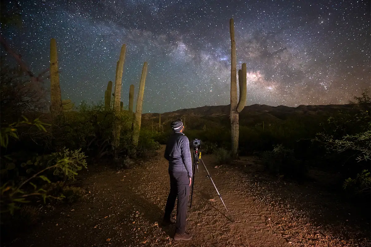 Photographer setting up a tripod for astrophotography under the Milky Way in a desert landscape.