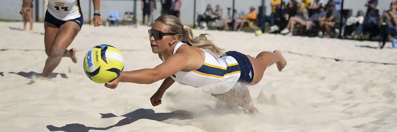 A beach volleyball player dives across the sand to reach the ball during a fast-moving sports action moment.