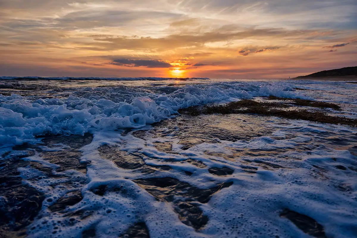 Ocean waves and sea foam fill the foreground during a warm golden-hour sunset.