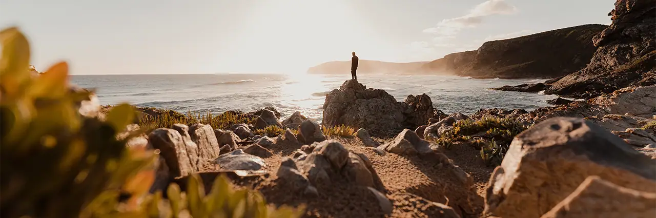 A person stands on coastal rocks during golden hour with the ocean and cliffs in the background.
