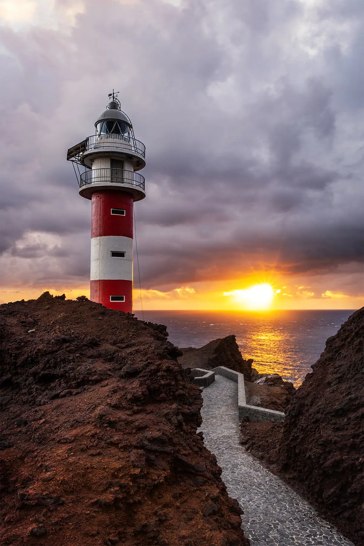 A stone path leads toward a red and white lighthouse at sunset beside the ocean.