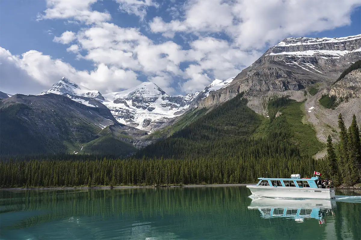 A boat crosses a turquoise mountain lake with snow-covered peaks and forest in the background.