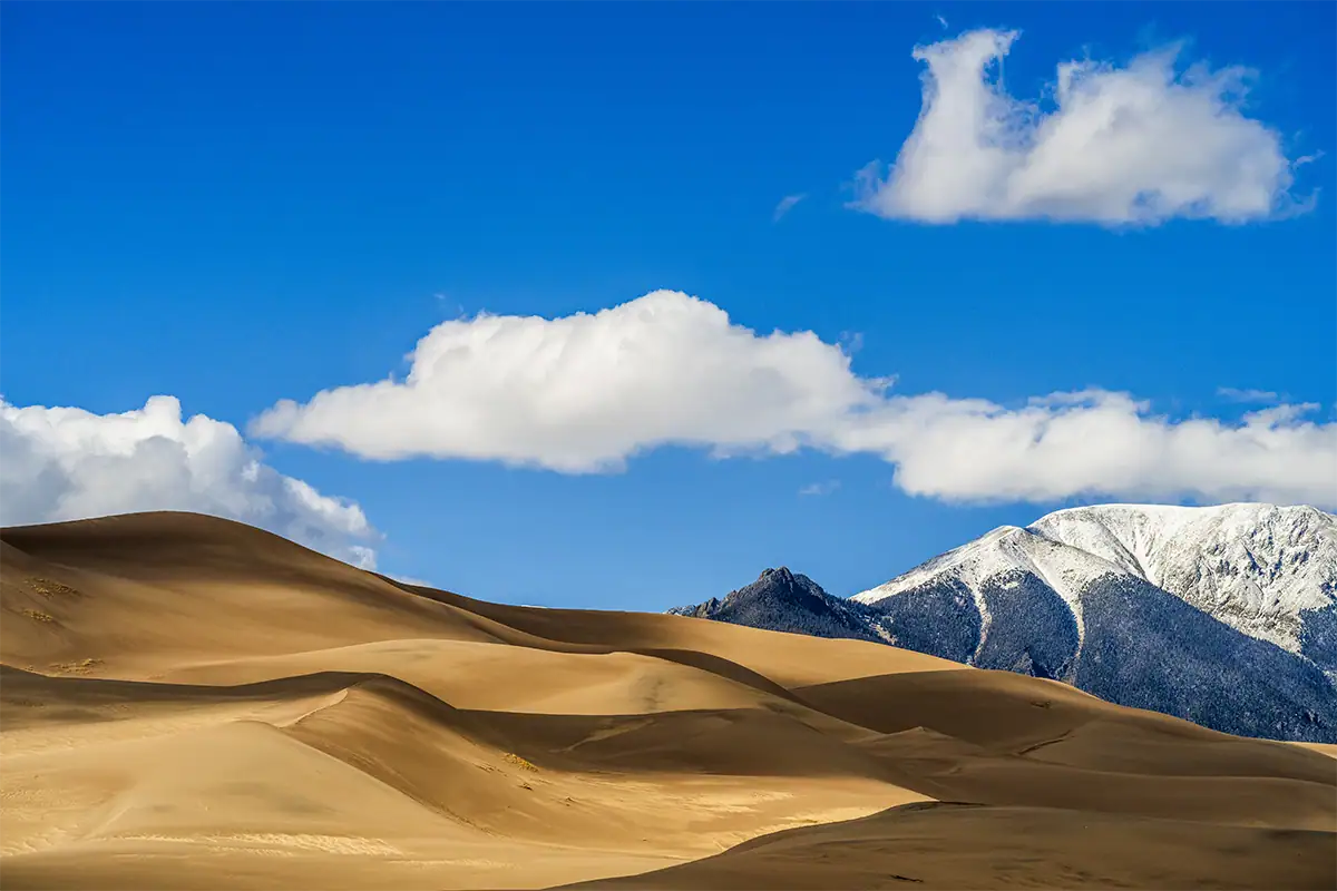 Golden sand dunes overlap with snow-covered mountains and clouds in the distance.