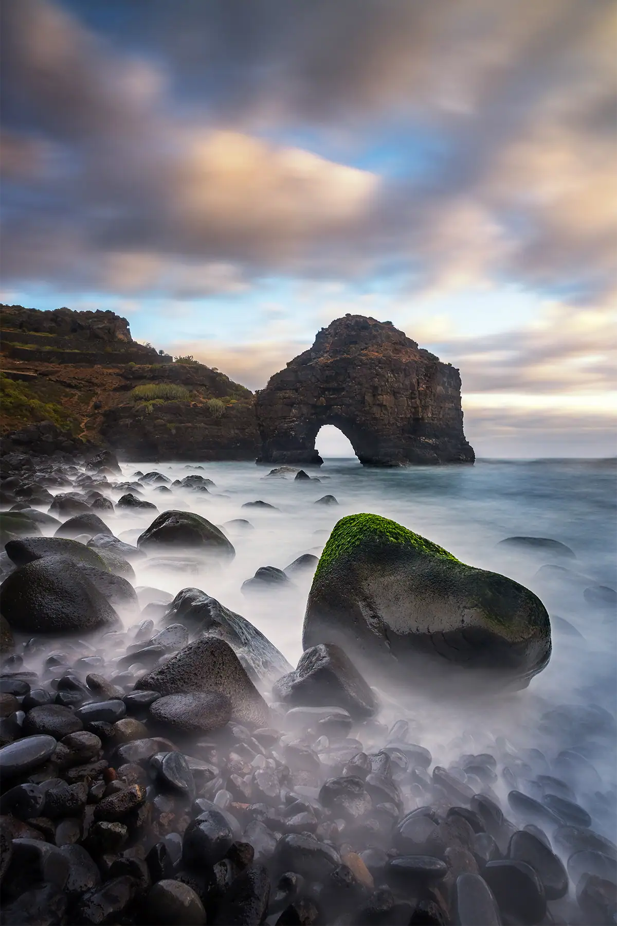 A rocky seascape with smooth long-exposure water, a natural arch, and dramatic clouds.