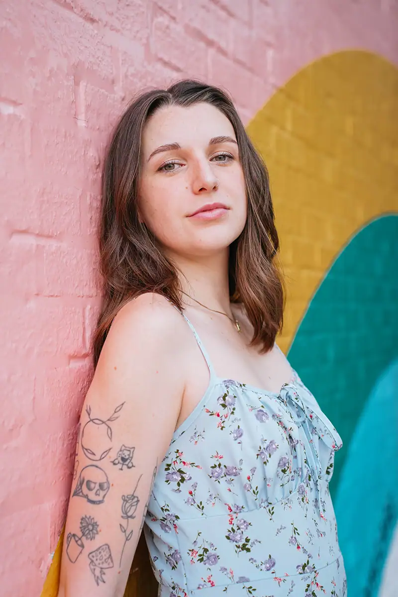 A woman with arm tattoos posed against a colorful wall mural.