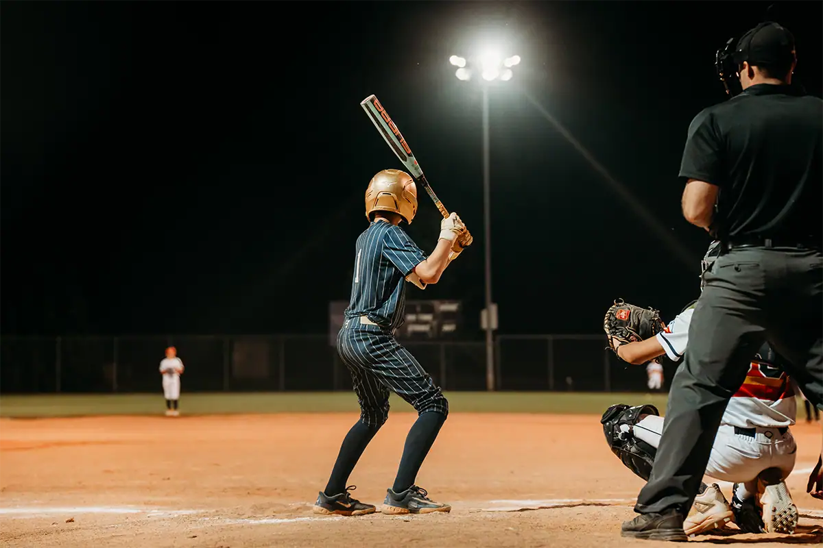 A youth baseball player bats during a night game under bright field lights.