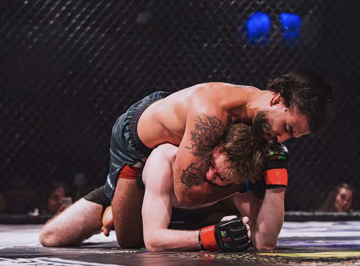 Low-light sports photography image of two MMA fighters grappling in an indoor arena.