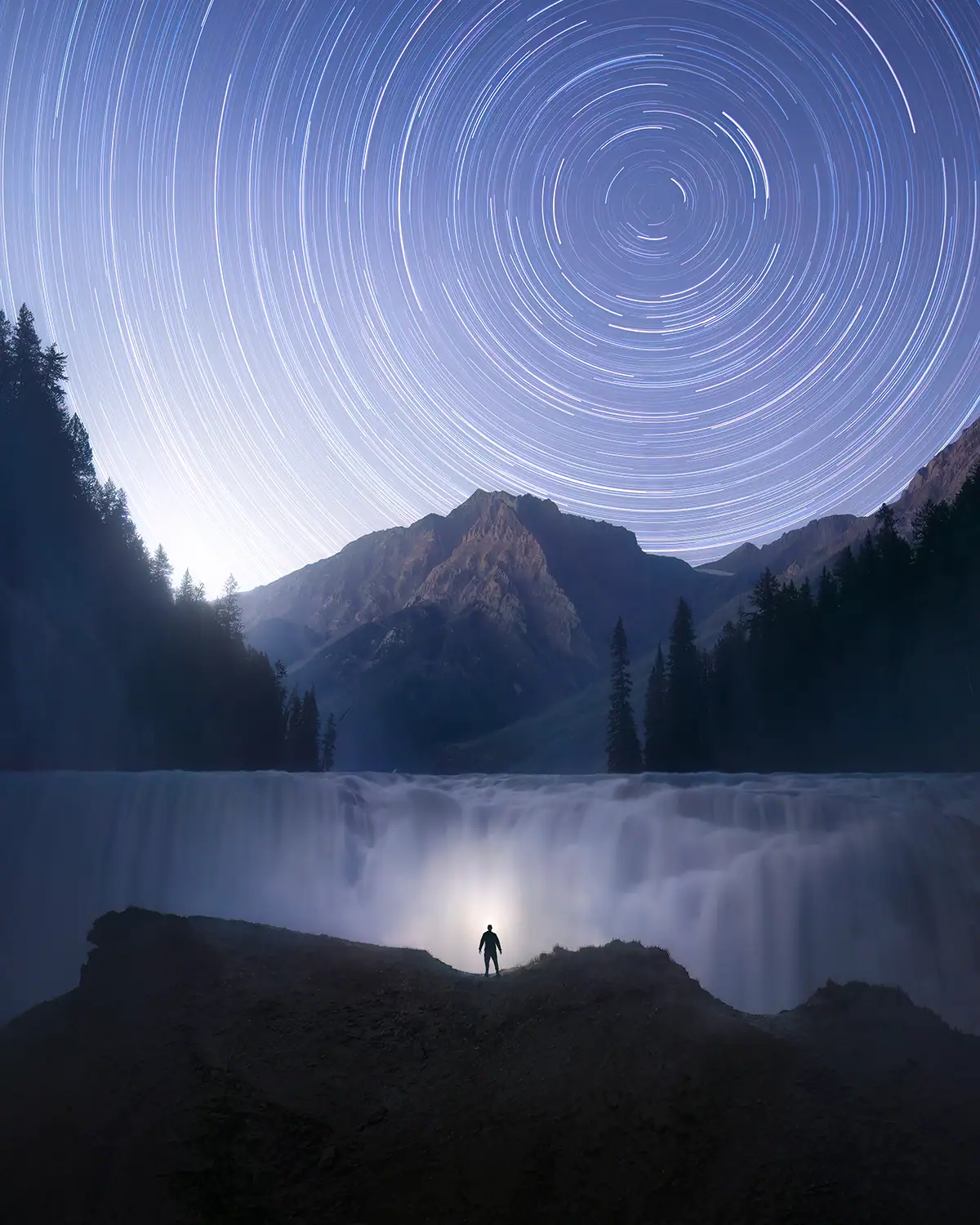 Stacked star trail photograph showing circular star arcs above a waterfall and mountain landscape.