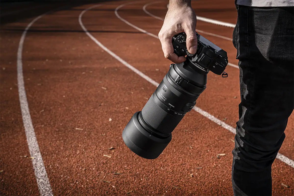 A photographer holds a Tamron 150-500mm lens beside a running track.