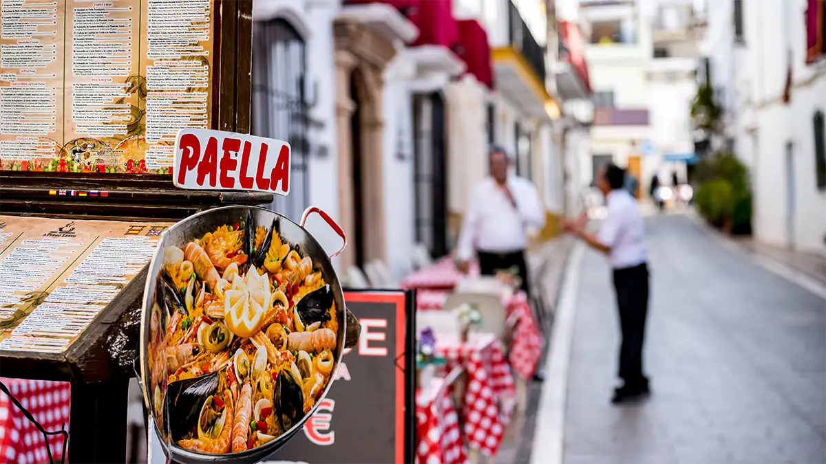 Paella sign on a Marbella side street with softly blurred restaurant staff in the background, photographed with the Tamron 35-100mm F2.8 travel photography lens