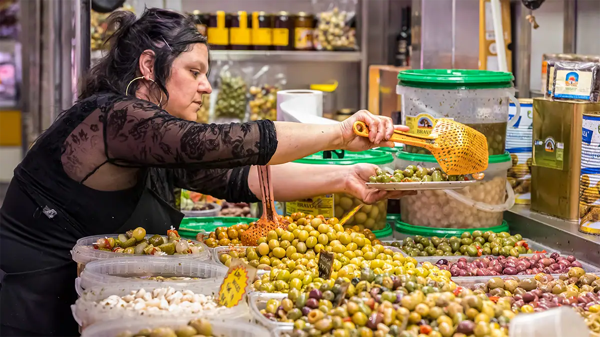 An olive vendor serves olives at a busy market in Spain, photographed with the Tamron 35-100mm F2.8 travel photography lens.