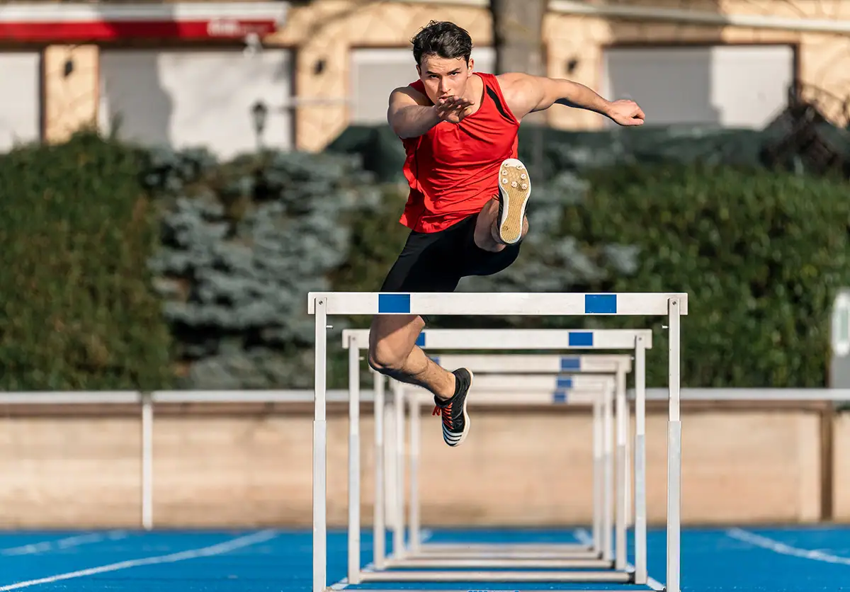 A hurdler clears a hurdle while running toward the camera in a track and field event.