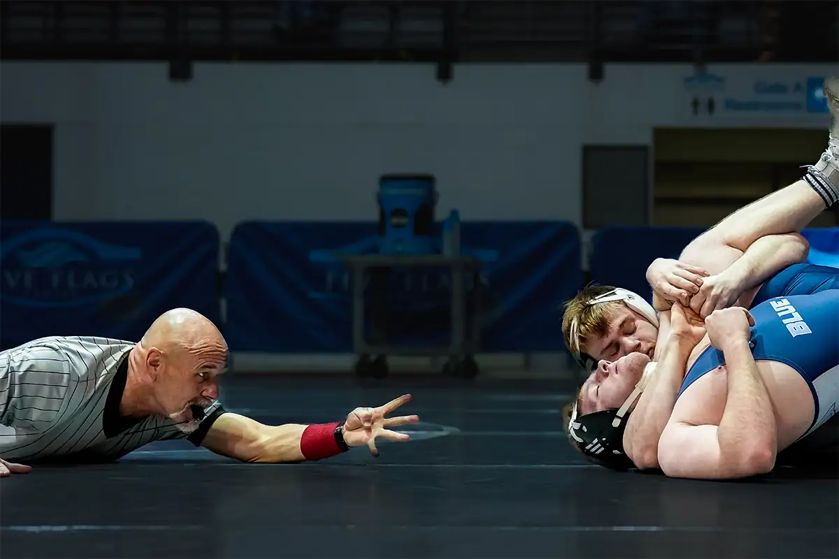 A referee watches two wrestlers compete from close range during an indoor sports match.