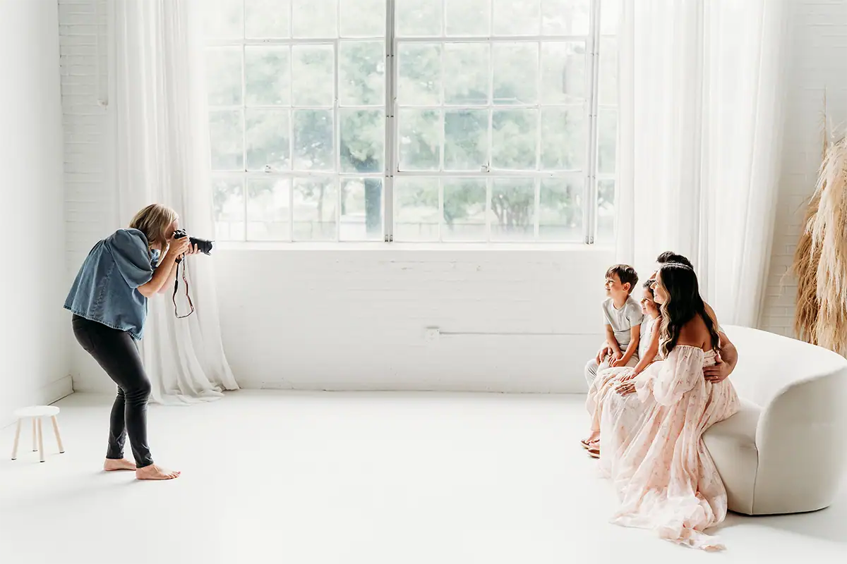 Fotógrafo tirando um retrato de família em um estúdio bem iluminado, com grandes janelas, onde a mãe e as duas crianças estão sentadas.