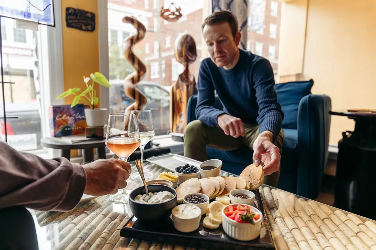 Un hombre que se dispone a tomar un bagel en la mesa de un café, donde hay vino, fruta y platillos de brunch, mientras el fondo aparece ligeramente desenfocado.