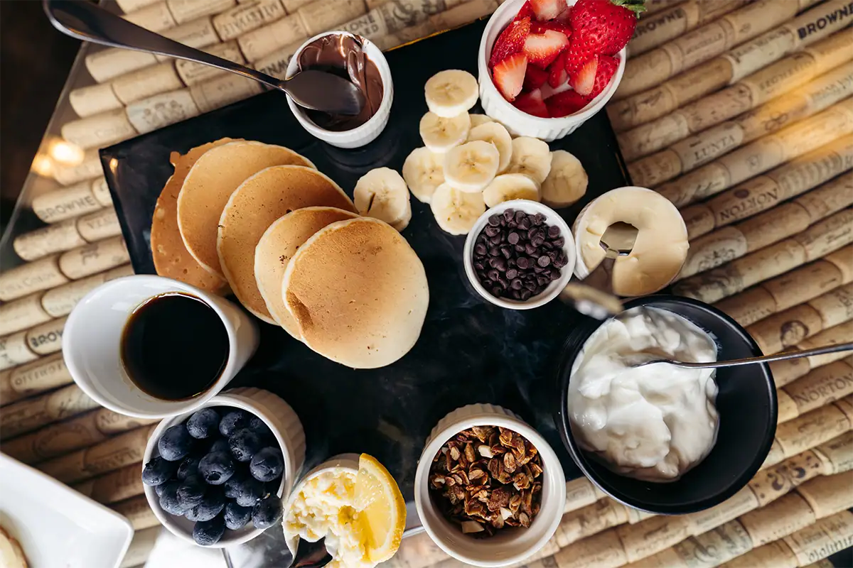 Foto tomada desde arriba de un café que muestra un brunch con panqueques, fruta, yogur, chocolate, granola y café sobre una mesa.
