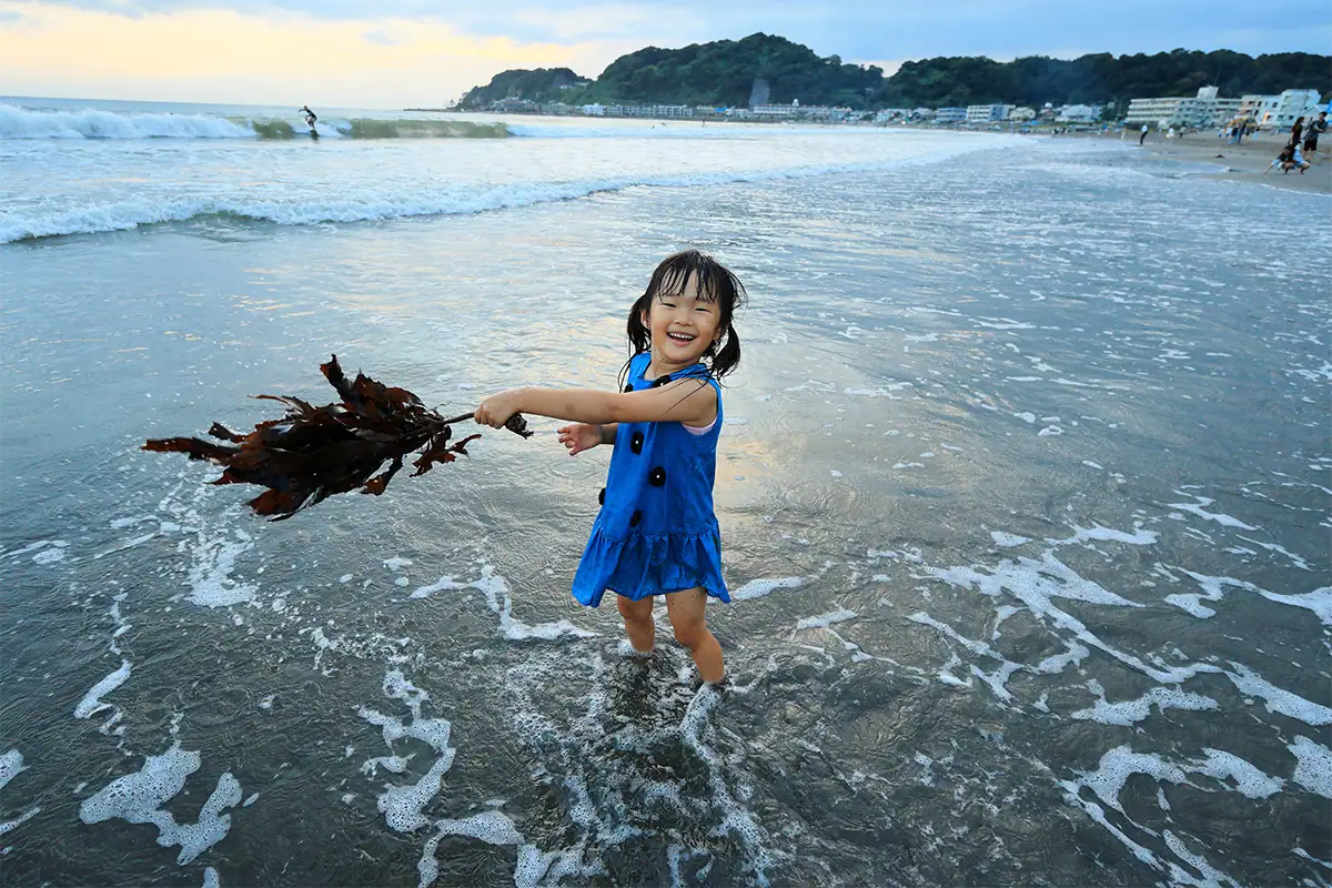 Menina vestindo um vestido azul, em pé na água rasa do mar, na praia, segurando algas marinhas e sorrindo enquanto as ondas quebram atrás dela.
