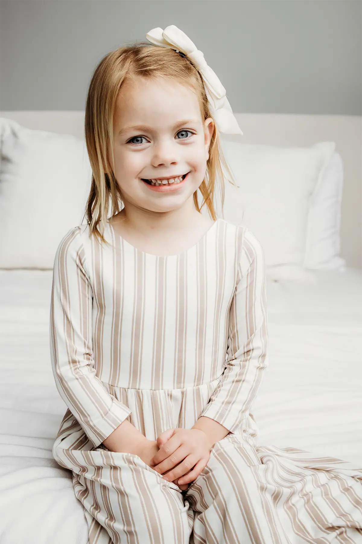 Menina sorridente, vestindo um vestido listrado e um laço branco, sentada em uma cama sob uma suave luz natural.