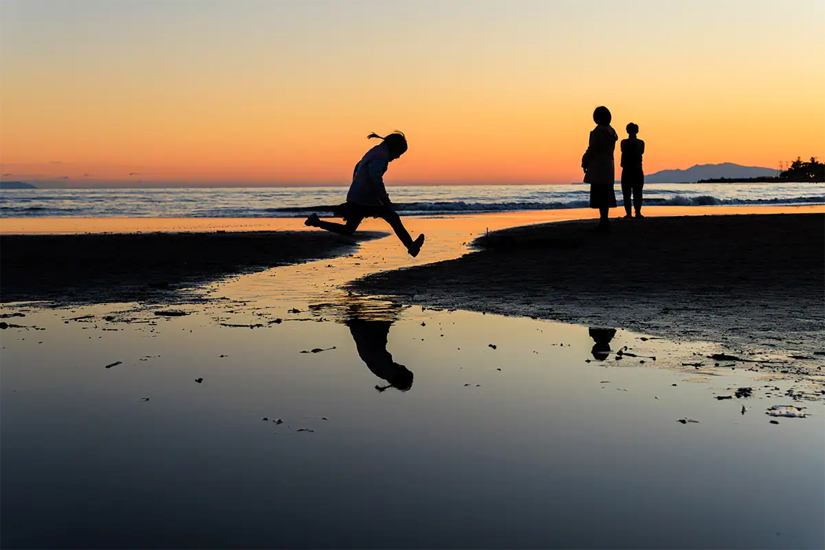 Silhouetted child jumping across a tidal stream at sunset as two people stand on the beach beside the ocean.