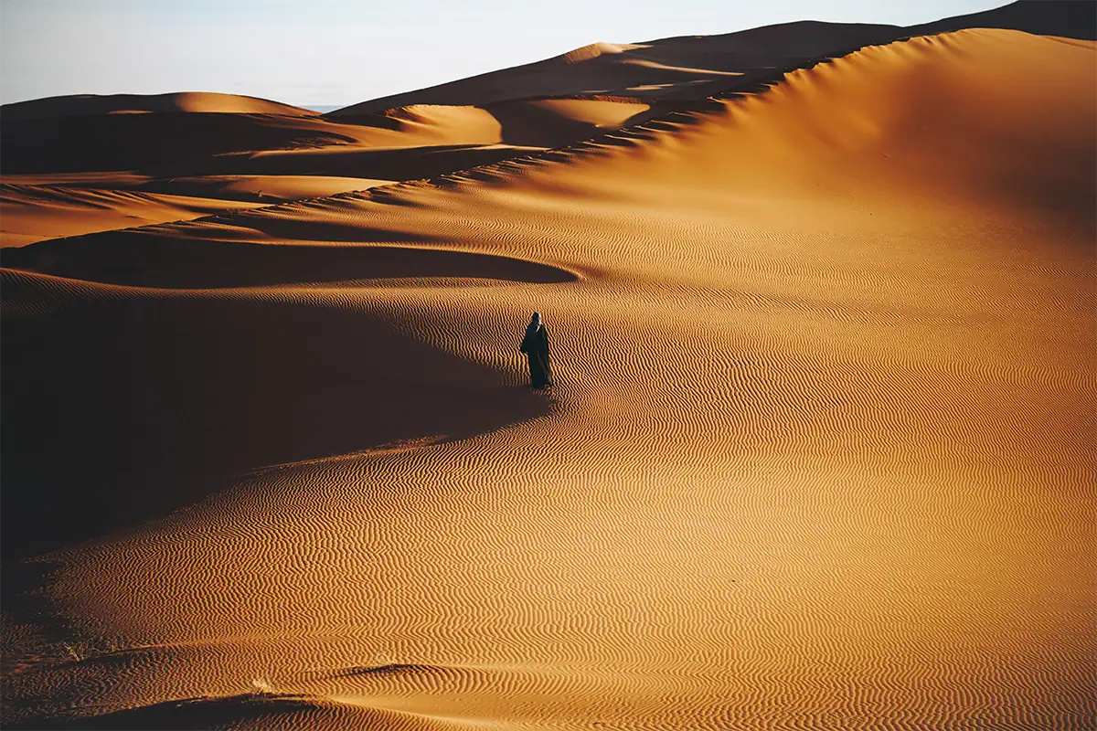 Lone traveler walking across golden desert dunes illuminated by warm, low-angle sunlight.