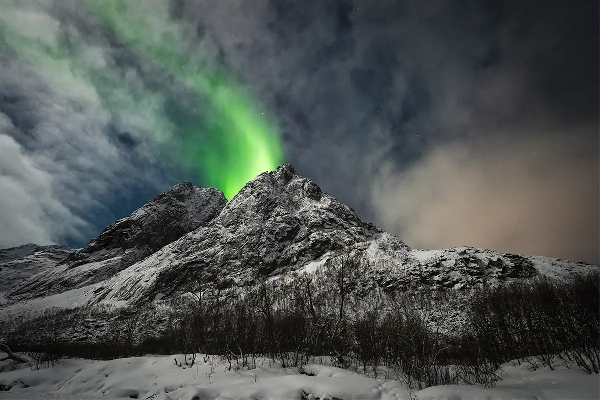 Aurora boreal fotografada sobre uma paisagem escura, com uma montanha coberta de neve e neve em primeiro plano.