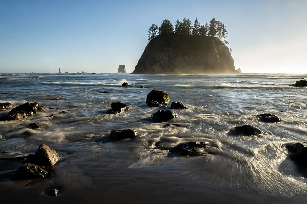 Golden-hour light illuminates ocean waves and rocky foreground beneath a forest-covered sea stack at Second Beach.