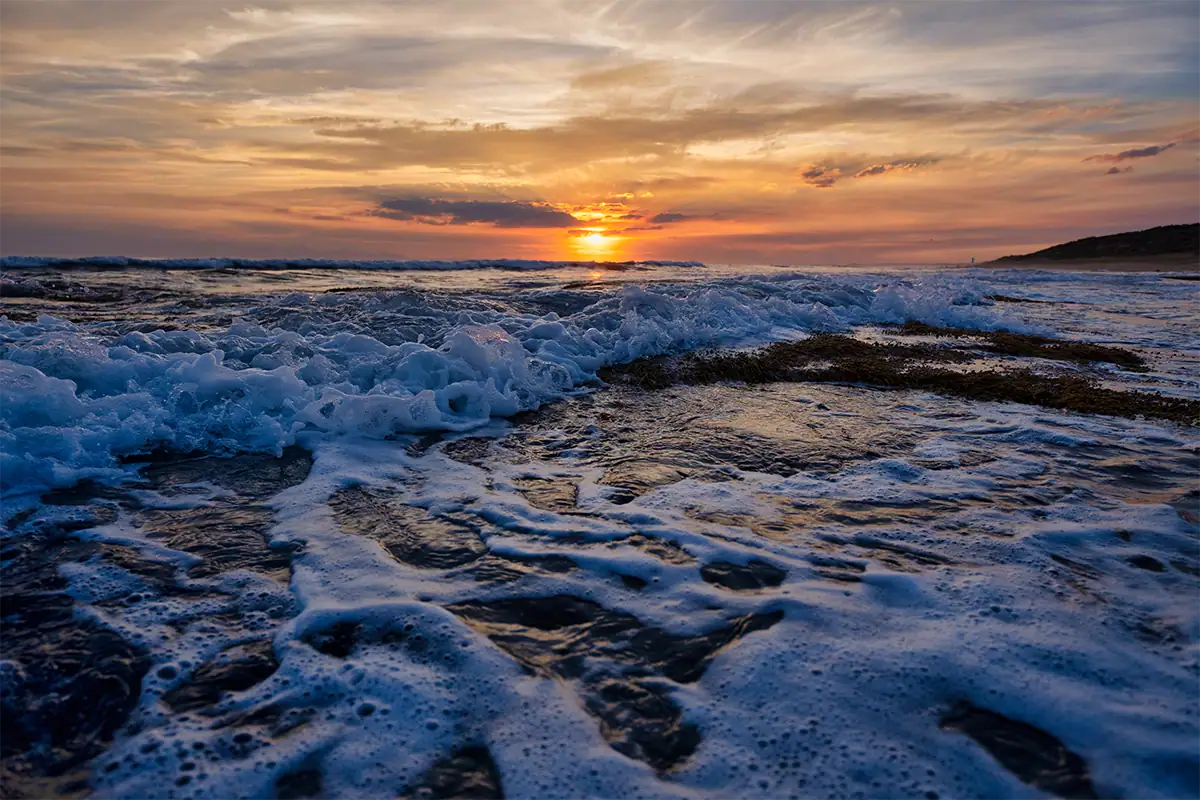 Sunrise above ocean waves washing across a beach beneath glowing orange and blue clouds.