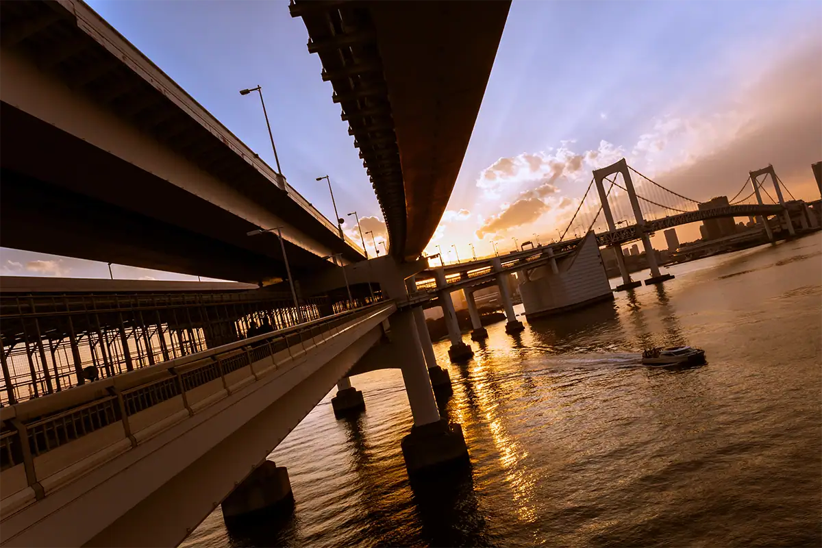 Golden sunset shining through bridge supports above a river as a boat passes beneath dramatic clouds.
