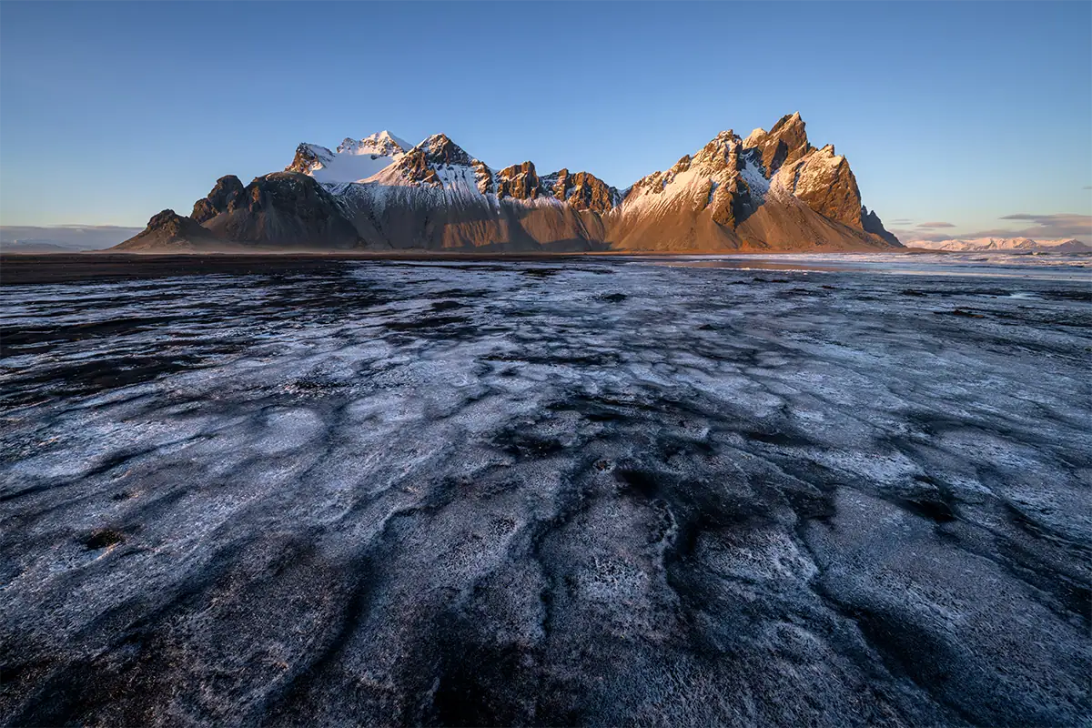 Golden hour light illuminates snow-covered mountains above a textured icy foreground in Iceland.