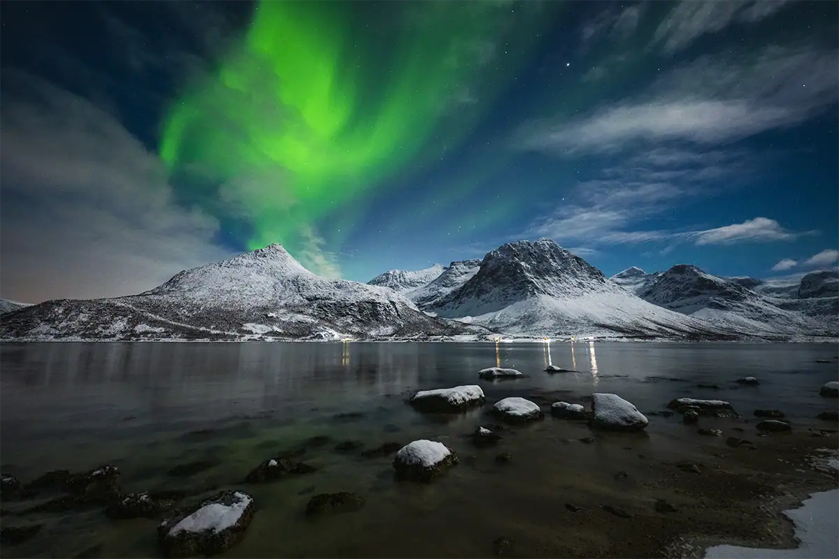 Northern lights glow above snow-covered mountains and a rocky shoreline in Norway, photographed with the Tamron 12-20mm F2.8.
