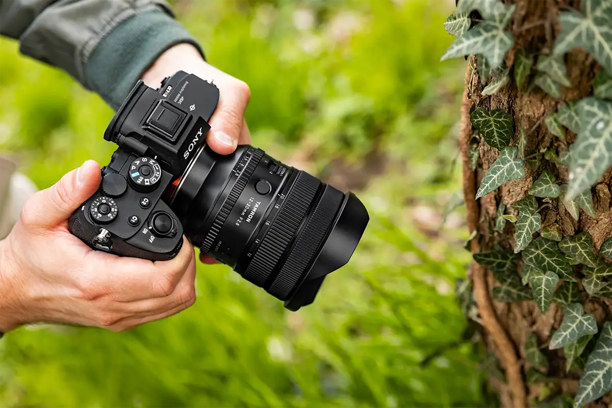 Photographer holding a Sony mirrorless camera with the Tamron 12-20mm F2.8 near an ivy-covered tree.